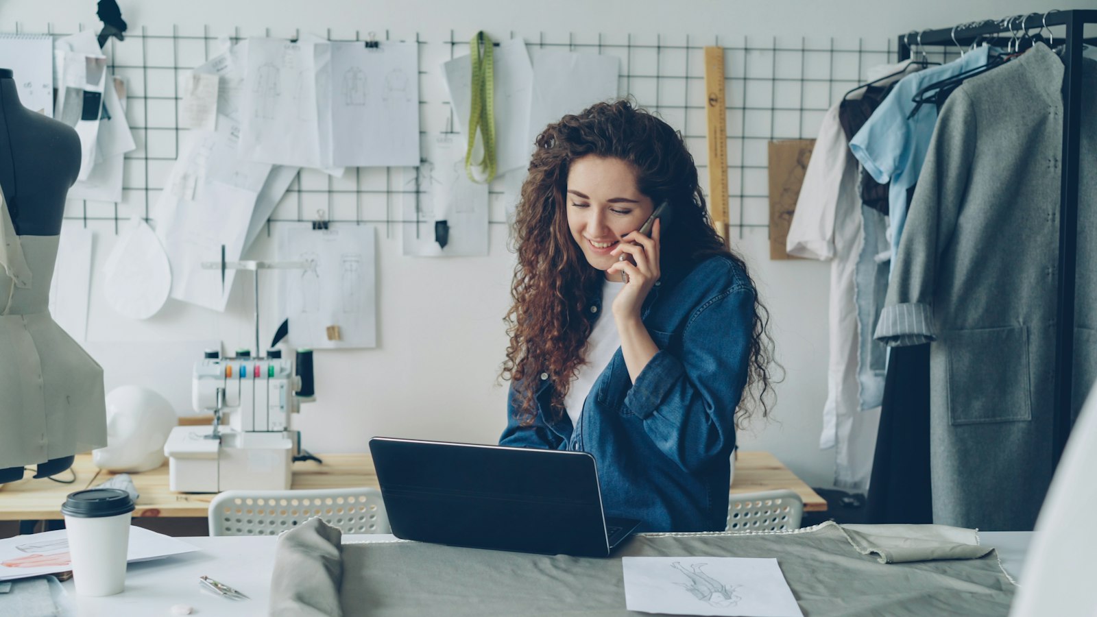 Small business owner in a tailoring studio, talking on the phone while working on a laptop